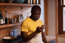 Vista lateral de cerca de un joven afroamericano con una camiseta amarilla de pie usando un teléfono inteligente en casa en su cocina - foto de stock