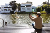 Vue de côté gros plan d'une femme caucasienne âgée assise au bord d'un lac et prenant des photos avec un smartphone — Photo de stock