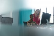 Side view of a young Caucasian woman standing and a young African American man sitting at a desk using a computer, talking and looking at the monitor together in the modern office of a creative business, with blurred foreground — Stock Photo