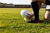 Visão lateral seção baixa de jogador de rugby feminino ajoelhado em um campo de rugby e definindo a bola em um tee para um chute de lugar — Fotografia de Stock