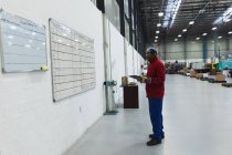 Vista lateral de un trabajador de una fábrica afroamericana de mediana edad sosteniendo un portapapeles comprobando la información en una pizarra blanca en un almacén en una planta de procesamiento - foto de stock