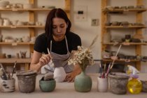 Vue de face gros plan d'une jeune potière caucasienne penchée sur une table de travail et vitrant un pot dans un atelier de poterie, avec des pots et des outils au premier plan — Photo de stock