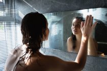 Por encima de la vista del hombro de una joven morena caucásica limpiando el vapor del espejo y mirándolo, de pie en un baño moderno - foto de stock