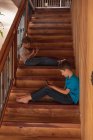 Vista lateral de dos niños caucásicos pre adolescentes sentados en una escalera en casa, utilizando teléfonos inteligentes - foto de stock