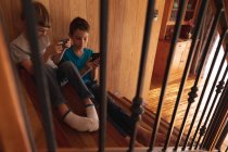 Вид спереди на двух кавказских мальчиков, сидящих дома на лестничной клетке с помощью смартфонов — стоковое фото