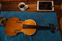 Vista ad alto angolo di un violino su un panno blu in un laboratorio di liuteria con tazza di caffè e tablet — Foto stock