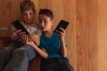 Vista frontal de cerca de dos niños caucásicos pre adolescentes sentados en una escalera en casa, utilizando teléfonos inteligentes - foto de stock