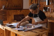 Vista lateral de una luthier hembra caucásica mayor midiendo en un banco de trabajo en su taller, con una tableta delante de ella y herramientas colgando en la pared en el fondo - foto de stock
