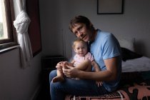 Retrato de un joven padre caucásico sosteniendo a su bebé, sentado en una cama - foto de stock