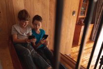 Vista frontal close-up de dois meninos pré-adolescentes caucasianos sentados em uma escada em casa, usando smartphones — Fotografia de Stock