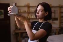 Side view close up of a young Caucasian female potter holding a clay flask and looking at it in a pottery studio — Stock Photo