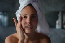 Ritratto da vicino di una giovane donna bruna caucasica con i capelli avvolti in un asciugamano, guardando dritto alla telecamera e toccandole il viso con una mano in un bagno moderno — Foto stock