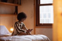 Вид збоку на молоду змішану жінку, яка використовує смартфон, сидячи на своєму ліжку вдома з приліжковою лампою, видно з дверного отвору — стокове фото