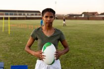 Retrato de uma jovem adulta de raça mista jogadora de rugby feminina em pé em um campo esportivo segurando uma bola de rugby durante uma sessão de treinamento — Fotografia de Stock