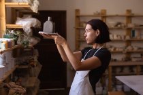 Vista lateral de una joven alfarera caucásica sosteniendo un matraz de barro y mirándolo en un estudio de cerámica - foto de stock
