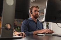 Vue de face gros plan d'un jeune homme caucasien assis à un bureau à l'aide d'un ordinateur et portant un casque dans un bureau créatif, vu entre les écrans d'ordinateur, avec la main d'un collègue travaillant à côté de lui sur le bureau — Photo de stock