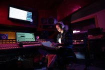 Вид сбоку на молодую кавказскую звукоинженершу, сидящую и работающую за микшерским столом в студии звукозаписи с помощью ноутбука и в наушниках — стоковое фото
