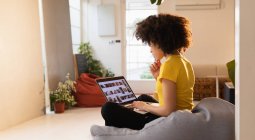 Вид сбоку молодой женщины смешанной расы, сидящей на бобовом мешке со скрещенными ногами с помощью ноутбука в креативном офисе — стоковое фото