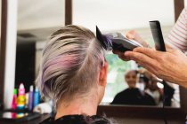 Visão traseira close-up de um cabeleireiro masculino caucasiano de meia idade e uma jovem mulher caucasiana ter seu cabelo aparado em um salão de cabeleireiro, refletido em um espelho — Fotografia de Stock