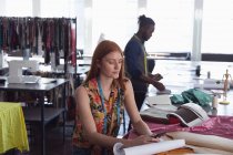 Vue de face d'une jeune étudiante de mode caucasienne travaillant sur un design dans un studio au collège de mode, avec un étudiant masculin travaillant en arrière-plan — Photo de stock