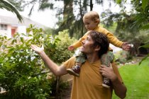 Front view of a young Caucasian father carrying his baby on his shoulders in a garden — Stock Photo