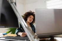 Вид спереду крупним планом молода змішана жінка, що сидить за столом, використовуючи ноутбук у творчому офісі — стокове фото