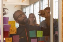 Передній погляд на молодого афроамериканця в окулярах пише нотатки на скляній стіні під час сесії командного мозкового штурму в творчому офісі, який видно через скляну стіну з колегами, які стоять позаду нього і дивляться — стокове фото
