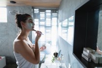 Vista lateral de uma jovem mulher morena caucasiana vestindo uma toalha de banho olhando no espelho segurando um frasco e aplicando um pacote de rosto em seu rosto com uma escova em um banheiro moderno — Fotografia de Stock
