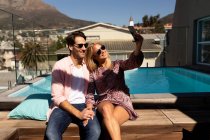 Vista frontal de una feliz pareja caucásica joven relajándose en vacaciones sentado junto a una piscina, tomando una selfie - foto de stock