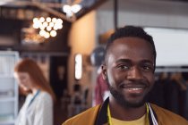 Портрет молодого афроамериканского студента-модельера, улыбающегося перед камерой в студии колледжа моды, на заднем плане которого работает студентка — стоковое фото
