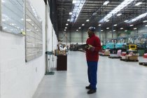 Vue latérale d'un ouvrier afro-américain d'âge moyen tenant un presse-papiers vérifiant les informations sur un tableau blanc dans un entrepôt d'une usine de transformation — Photo de stock