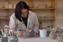 Vue de face gros plan d'une jeune potière caucasienne assise à une table de travail peignant une glaçure colorée sur une fiole d'argile dans un atelier de poterie — Photo de stock