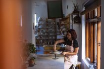 Вид збоку молодого Кавказького жіночого Поттера сидить за робочим столом перед вікном, працюючи з глиною на обв'язці коліс в гончарному студії — стокове фото