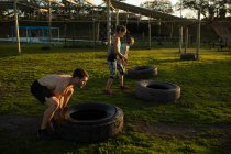 Vista lateral de duas jovens mulheres caucasianas e um jovem homem caucasiano virando pneus em um ginásio ao ar livre durante uma sessão de treinamento de bootcamp — Fotografia de Stock