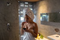 Vorderansicht einer jungen kaukasischen Frau, deren Haare in ein Handtuch gehüllt sind, sich die Zähne putzt und in einem modernen Badezimmer auf ihr Smartphone blickt — Stockfoto