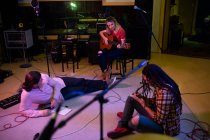 Vista frontal de una joven mujer caucásica sentada en un taburete tocando una guitarra acústica mientras un joven hombre caucásico yace en el suelo escuchándola y un joven mestizo sentado en el suelo escribiendo - foto de stock
