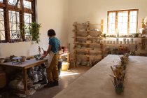 Vue arrière d'une jeune potière caucasienne travaillant avec des morceaux d'argile debout à une table de travail devant une fenêtre dans un atelier de poterie — Photo de stock