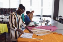Вид сбоку на молодого афроамериканского студента-модельера, работающего над дизайном в студии колледжа моды, со студентками, работающими на заднем плане — стоковое фото