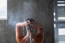 Close up visão traseira de uma jovem mulher morena caucasiana de pé sob o chuveiro shampooing seu cabelo em um banheiro moderno — Fotografia de Stock