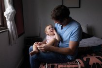 Front view of a young Caucasian father holding his baby, sitting on a bed and looking at each other — Stock Photo