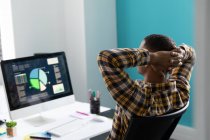 Vue arrière gros plan d'un jeune Afro-Américain assis à un bureau les mains derrière la tête, regardant un moniteur d'ordinateur dans le bureau moderne d'une entreprise créative — Photo de stock