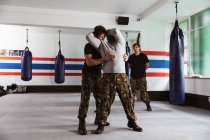 Vue de face d'un instructeur caucasien d'âge moyen qui donne un entraînement d'autodéfense dans un gymnase de boxe démontrant son emprise sur un jeune homme de race mixte, tandis qu'un autre jeune homme regarde — Photo de stock