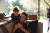 Vista frontal de perto de dois meninos pré-adolescentes usando um smartphone e ouvindo música com fones de ouvido em uma sala de estar em casa — Fotografia de Stock