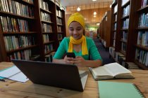 Вид спереди на молодую азиатскую студентку в хиджабе, держащую смартфон с помощью ноутбука и занимающуюся в библиотеке — стоковое фото