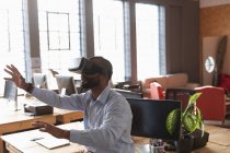 Side view close up of a young African American man sitting on a desk wearing a VR headset with his arms outstretched and hands out in a creative office — Stock Photo