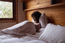 Vista lateral de uma jovem mulher de raça mista sentada na cama apoiada em um travesseiro usando um smartphone em casa — Fotografia de Stock