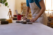 Vue latérale section médiane d'une potière portant un tablier travaillant avec des morceaux d'argile à une table de travail dans un atelier de poterie — Photo de stock