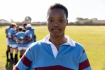 Porträt einer jungen erwachsenen gemischten Rugby-Spielerin, die auf einem Rugbyfeld steht und in die Kamera blickt, während sich ihre Teamkolleginnen im Hintergrund eng aneinander drängen — Stockfoto