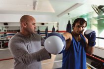 Front view close up of a young mixed race male boxer punching being instructed by a middle aged Caucasian male trainer in a boxing gym — Stock Photo