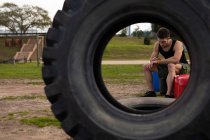 Vista frontal de um jovem caucasiano sentado e checando seu relógio em um ginásio ao ar livre antes de uma sessão de treinamento de bootcamp, visto através de um pneu — Fotografia de Stock